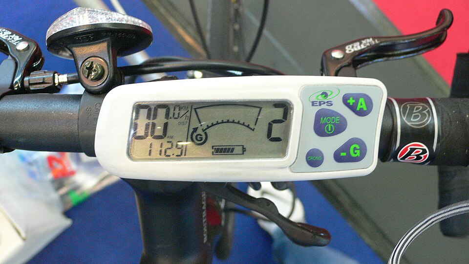 An e-bike handlebar display showing speed, assist level and a battery bar, with buttons beside it